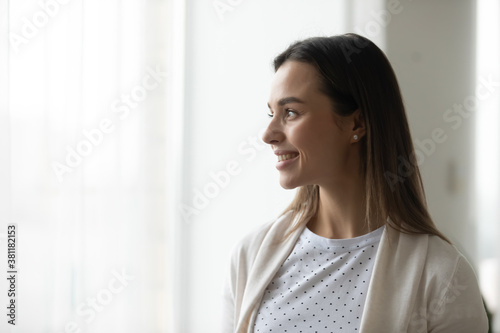 Smiling millennial Caucasian girl look in window distance at home feel optimistic dreaming or visualizing. Happy thoughtful dreamy young woman lost in thoughts, planning or imagining. Vision concept.