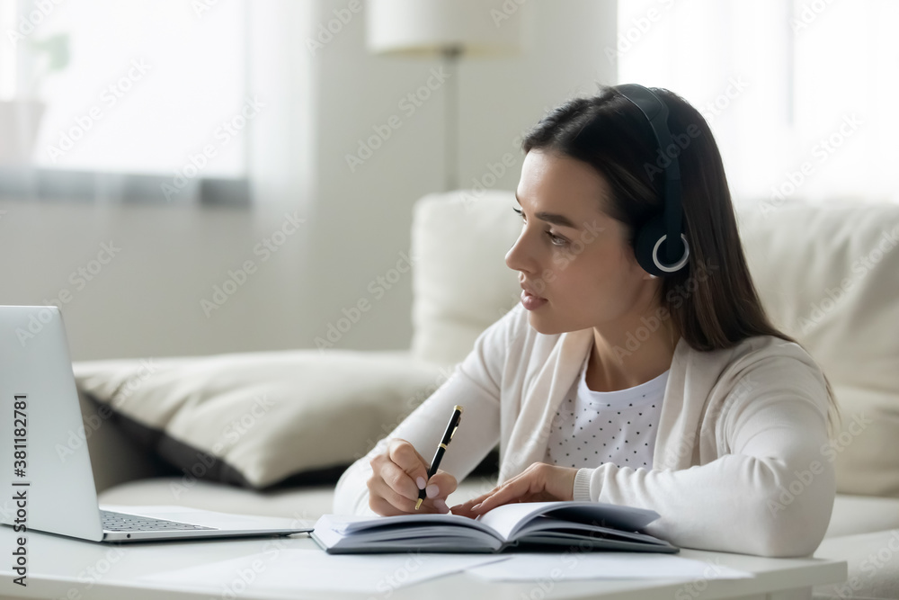 Focused millennial female student in headphones look at laptop screen take online study course or training. Young woman in earphones watch webinar on computer make notes. Distant education concept. Focused millennial female student in headphones look at laptop screen take online study course or training. Young woman in earphones watch webinar on computer make notes. Distant education concept.