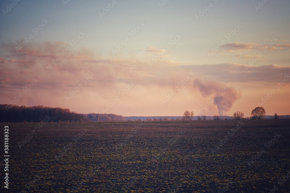 Smoke from a large fire over an agricultural field in the evening. Puffs of smoke in the sunset sky.