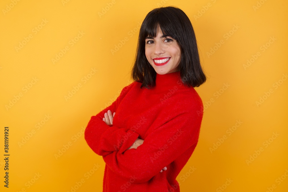 Caucasian brunette woman wearing red casual sweater isolated over yellow background crossing arms looking aside having a nice conversation