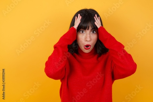 Caucasian brunette woman wearing red casual sweater isolated over yellow background with scared expression, keeps hands on head, jaw dropped, has terrific expression. Omg concept © Roquillo