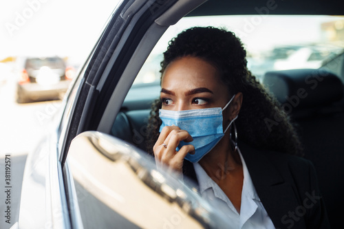 Young businesswoman being a taxi passenger and having a ride wearing and adjusting medical mask for health protection. Business trips during pandemic, new normal and coronavirus travel safety concept.