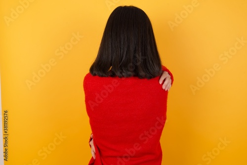 Caucasian brunette woman wearing red casual sweater isolated over yellow background hugging herself happy and positive from backwards. Self love and self care