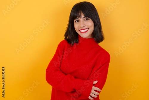 Caucasian brunette woman wearing red casual sweater isolated over yellow background happy face smiling with crossed arms looking at the camera. Positive person. © Roquillo
