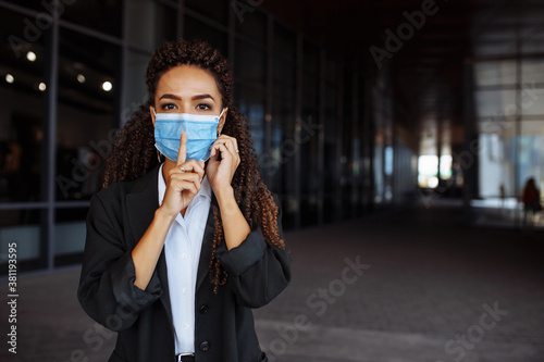 Young businesswoman wearing a medical mask shows silence sign with her finger near the mouth while talking on the mobile phone. Lack of information during Covid-19 pandemic concept.