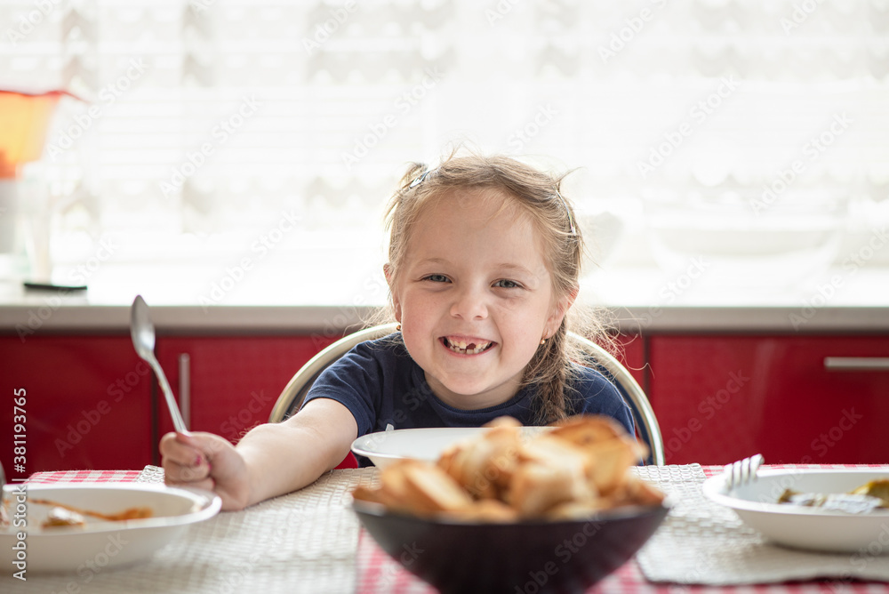 Toothless and happy six years old caucasian girl smiling. Adorable child sit near the table with spoon in hands and laughing, home kitchen Toothless and happy six years old caucasian girl smiling. Adorable child sit near the table with spoon in hands and laughing, home kitchen