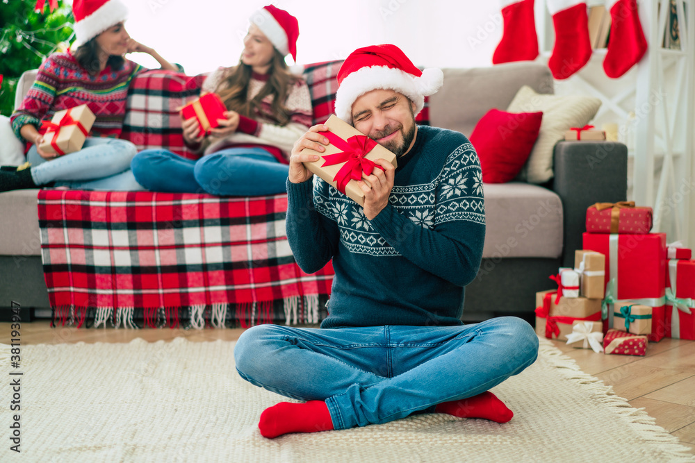 Obraz premium Young happy handsome bearded man in a Santa hat and winter sweater is posing with a gift box in hands and showing on camera with smiling friends on the background.
