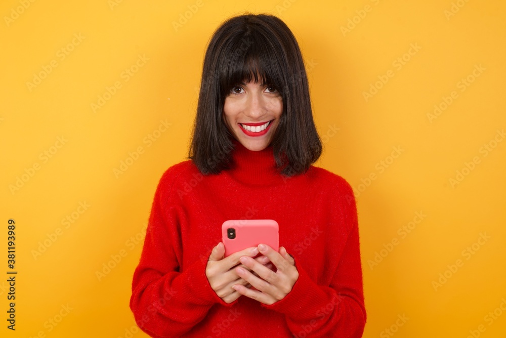 Caucasian brunette woman wearing red casual sweater enjoys distant communication, uses mobile phone, surfs fast unlimited internet, has pleasant smile, makes shopping online, isolated on yellow Caucasian brunette woman wearing red casual sweater enjoys distant communication, uses mobile phone, surfs fast unlimited internet, has pleasant smile, makes shopping online, isolated on yellow