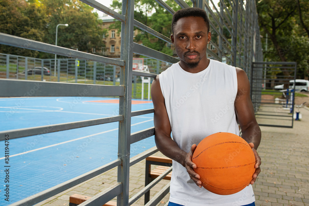 A man in a white T-shirt holds a basketball in his hands in front of him, looking straight at the playground. The concept of controversy, healthy living