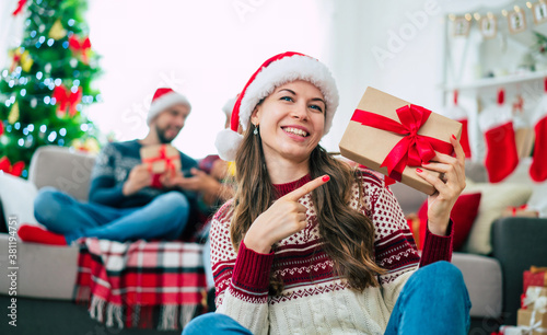 Beautiful young happy smiling woman in a Christmas sweater and Santa hat is holding a colorful gift box in hands at home on the background of her friends and Christmas tree.