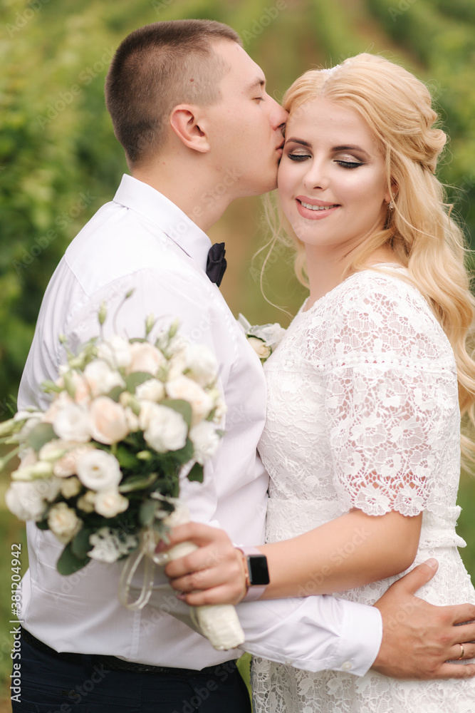 Groom and bride walking in vineyard in their wedding day. Blond hair woman in white dress hold wedding bouquet in hands Groom and bride walking in vineyard in their wedding day. Blond hair woman in white dress hold wedding bouquet in hands