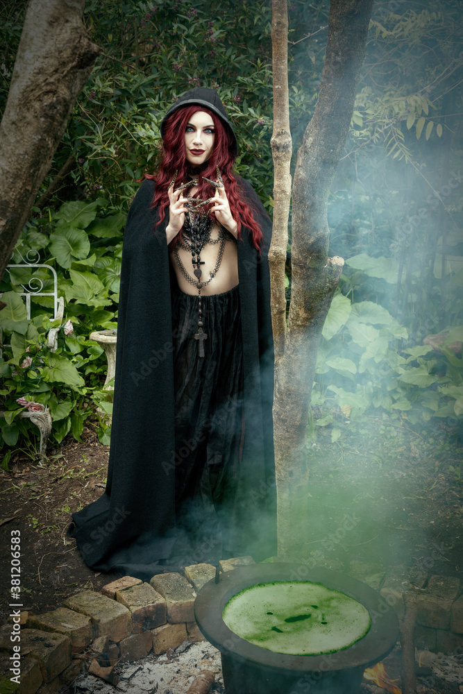 Woman wearing a black cape stands in a Forest. The image has a Halloween theme.