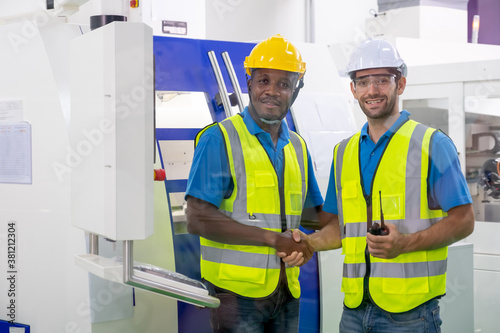 Soft blur of two factory worker men with uniform shaking hands also smiling and look to camera in front of machine. Concept of good management system to support industrial business working.