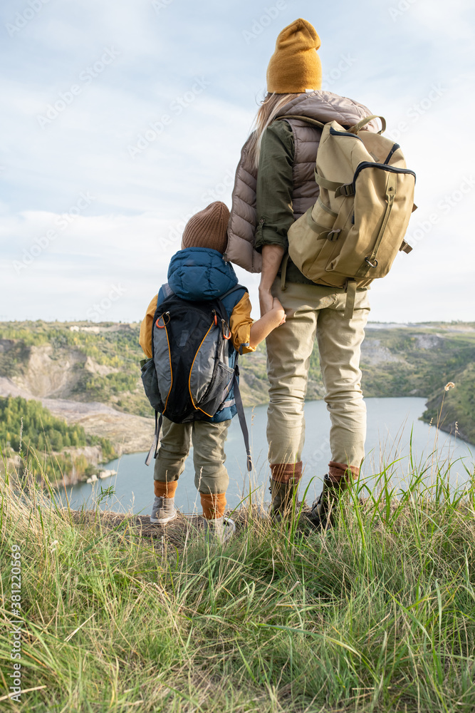 Rear view of young mother and little son with backpacks standing by lake Rear view of young mother and little son with backpacks standing by lake