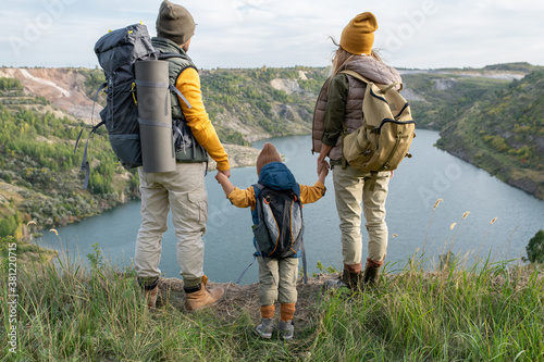 Rear view of young parents and their son with backpacks looking at lake or river