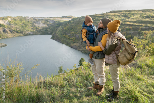 Young parents and their little son with backpacks standing against lake or river