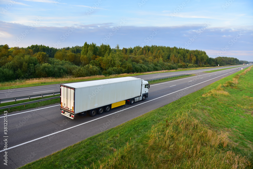 White truck with semi-trailer driving along highway on the sunset background. Goods delivery by roads. Services and Transport logistics. Object in motion, soft focus.