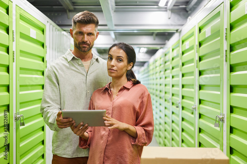 Waist up portrait of beautiful modem couple holding digital tablet while standing by self storage unit and looking at camera, copy space photo