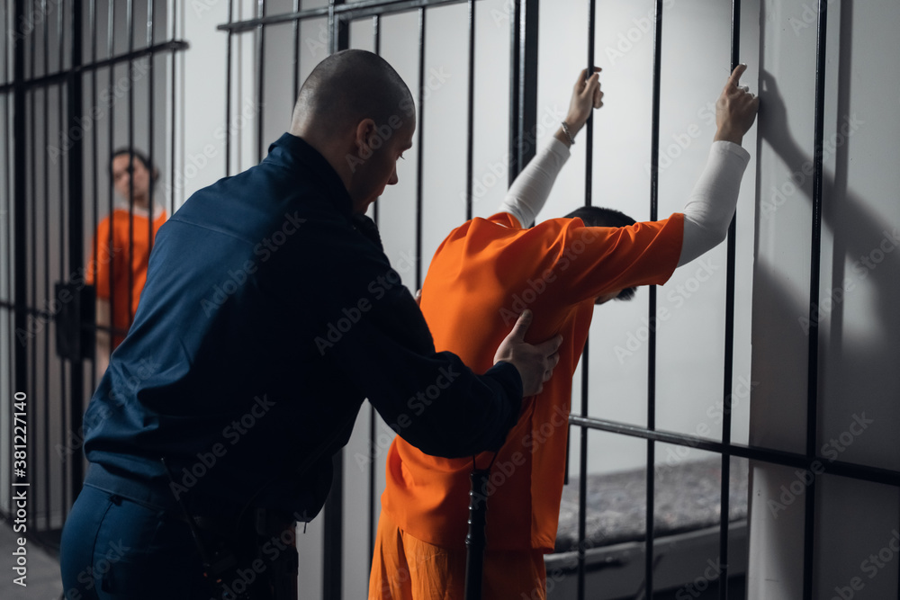 An armed guard searches a newly arrived criminal in a prison corridor against a backdrop of bars An armed guard searches a newly arrived criminal in a prison corridor against a backdrop of bars