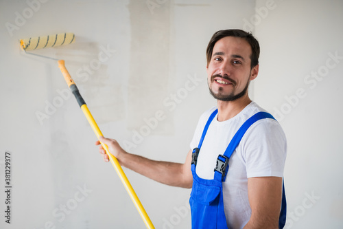 A bearded workman in blue uniform paints the wall with a roller. He smiles friendly and poses for the camera