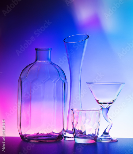 Still life with four backlit glass objects on a bright color gradient background in blue and pink tones. Vase, two glasses and a large pot.