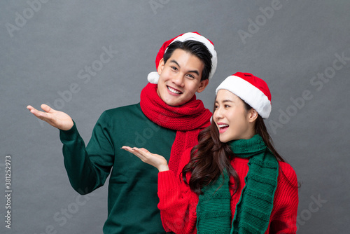 Surprised smiling Asian couple in Christmas attire doing open hand gesture for product placement on gray studio background