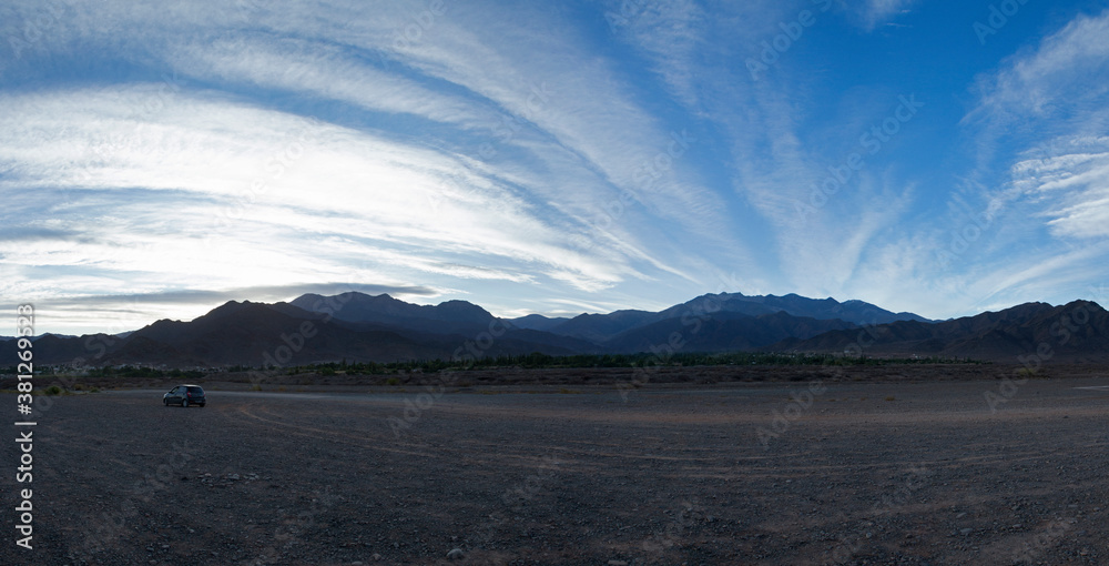Travel. Panorama view of a car parked in the dirt road across the desert and mountains under a beautiful sunrise blue sky. 