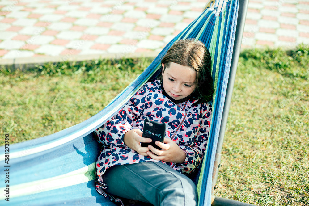 Cute caucasian girl 7-8 years old lies on hammock and using her phone on a sunny day Cute caucasian girl 7-8 years old lies on hammock and using her phone on a sunny day
