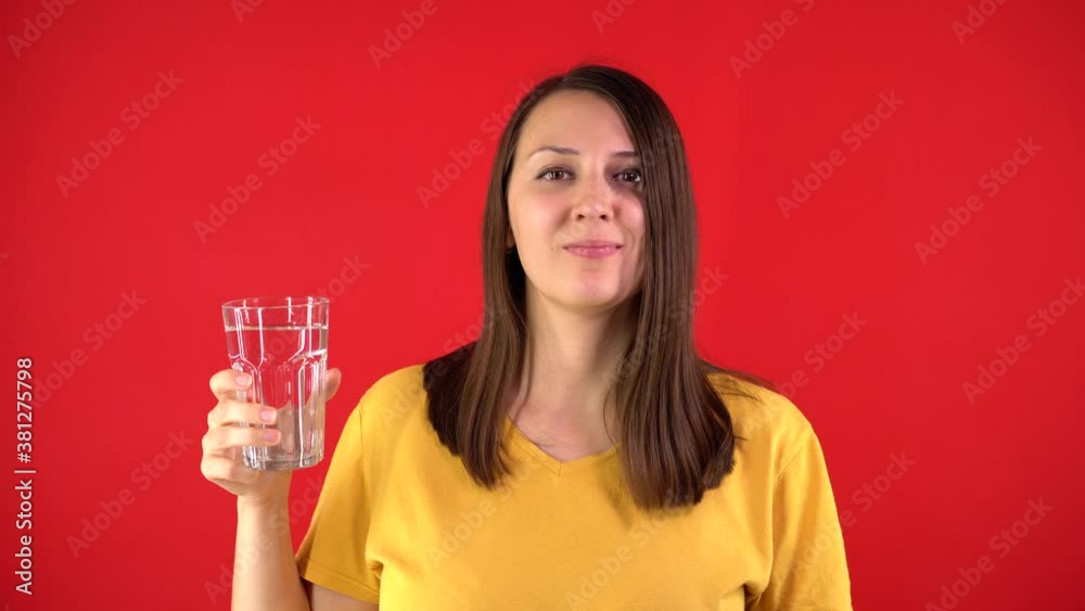 Attractive girl in a yellow T-shirt drinks water from a glass and shows a super sign on a red background. Healthy lifestyle concept. Beautiful girl holding a glass of drinking water, smiling. Attractive girl in a yellow T-shirt drinks water from a glass and shows a super sign on a red background. Healthy lifestyle concept. Beautiful girl holding a glass of drinking water, smiling.