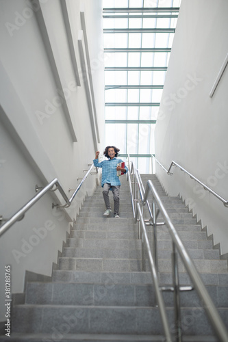 Schoolboy raising his hand, running downstairs with pile of books