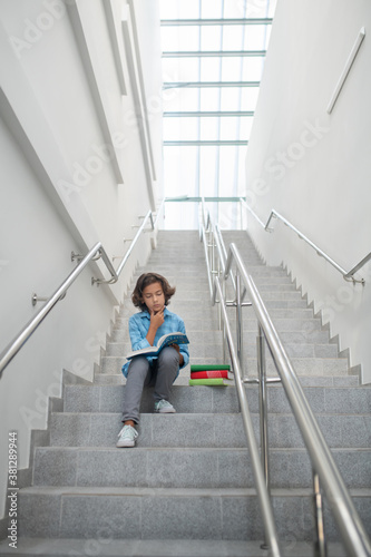 Schoolboy sitting on stairs, reading book, holding his chin