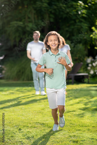 Excited boy running on lawn, his parents walking behind