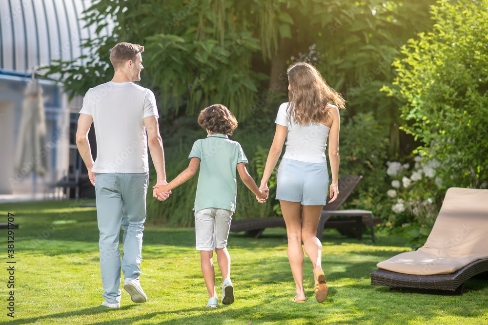 Fototapeta premium Back view of family family of three holding hands, walking on grass