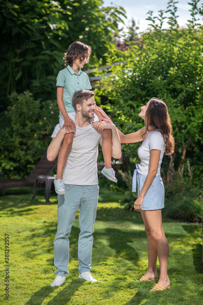 Fototapeta premium Father holding son on shoulders, his wife touching his hand, looking at them