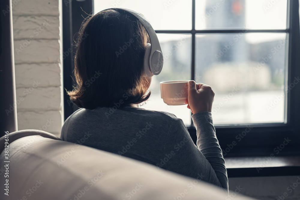 Back view of lonely сaucasian young woman enjoying having breakfast with cup of coffee and listenning music in headphones sitting near window at cold winter morning. Back view of lonely сaucasian young woman enjoying having breakfast with cup of coffee and listenning music in headphones sitting near window at cold winter morning.