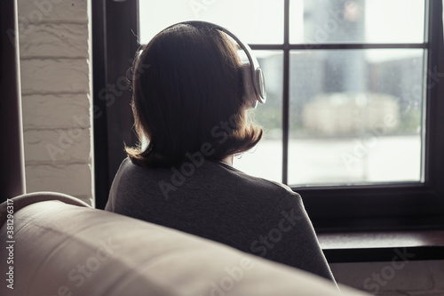 Back view of сaucasian young woman in headphones enjoying listening music sitting near window of apartments.