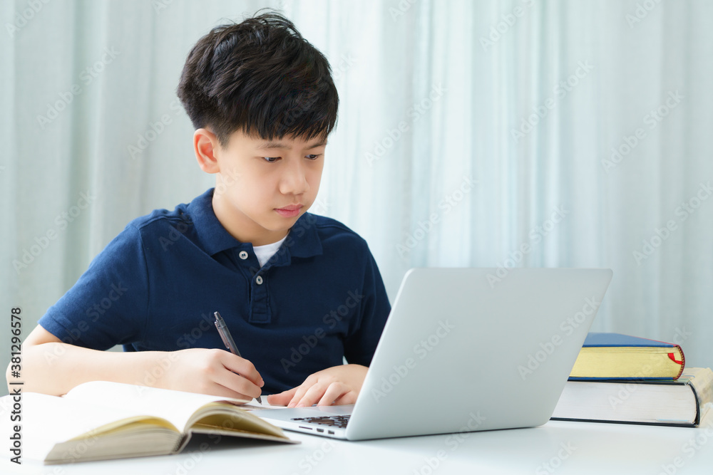 A smart and handsome asian teenager boy using notebook computer at the table to attend online classroom he look at screen and take notes while studying. Covid-19 New normal, Home Based Learning. A smart and handsome asian teenager boy using notebook computer at the table to attend online classroom he look at screen and take notes while studying. Covid-19 New normal, Home Based Learning.