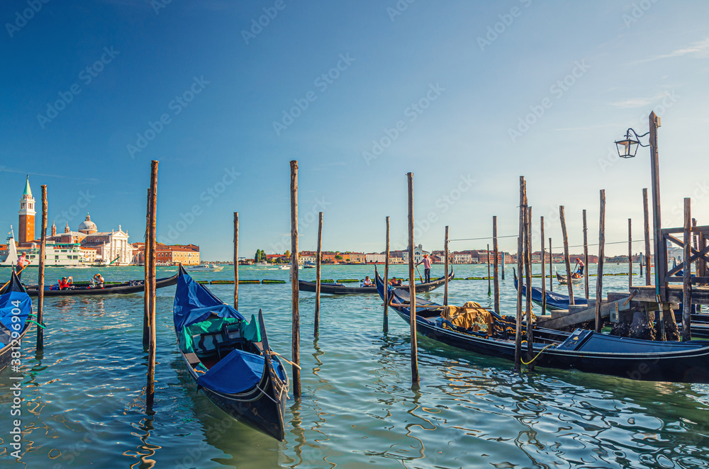 Gondolas moored docked on water in Venice city. Gondoliers sailing in San Marco basin waterway. San Giorgio Maggiore island with Campanile San Giorgio in Venetian Lagoon and Giudecca island, Italy