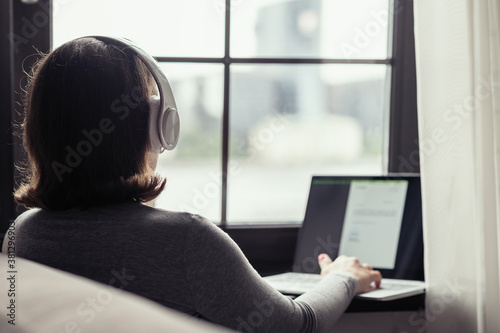 Back view of lonely woman listenning music in headphones and working on laptop sitting near window in cafe.