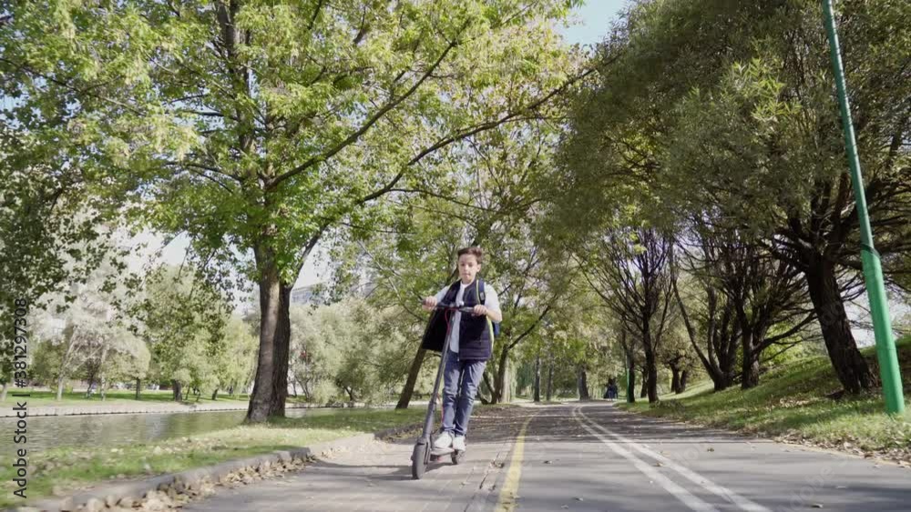School boy riding with his electric scooter in the city with backpack on sunny day. Child in colorful clothes biking on way to school. Electric transport. School boy riding with his electric scooter in the city with backpack on sunny day. Child in colorful clothes biking on way to school. Electric transport.