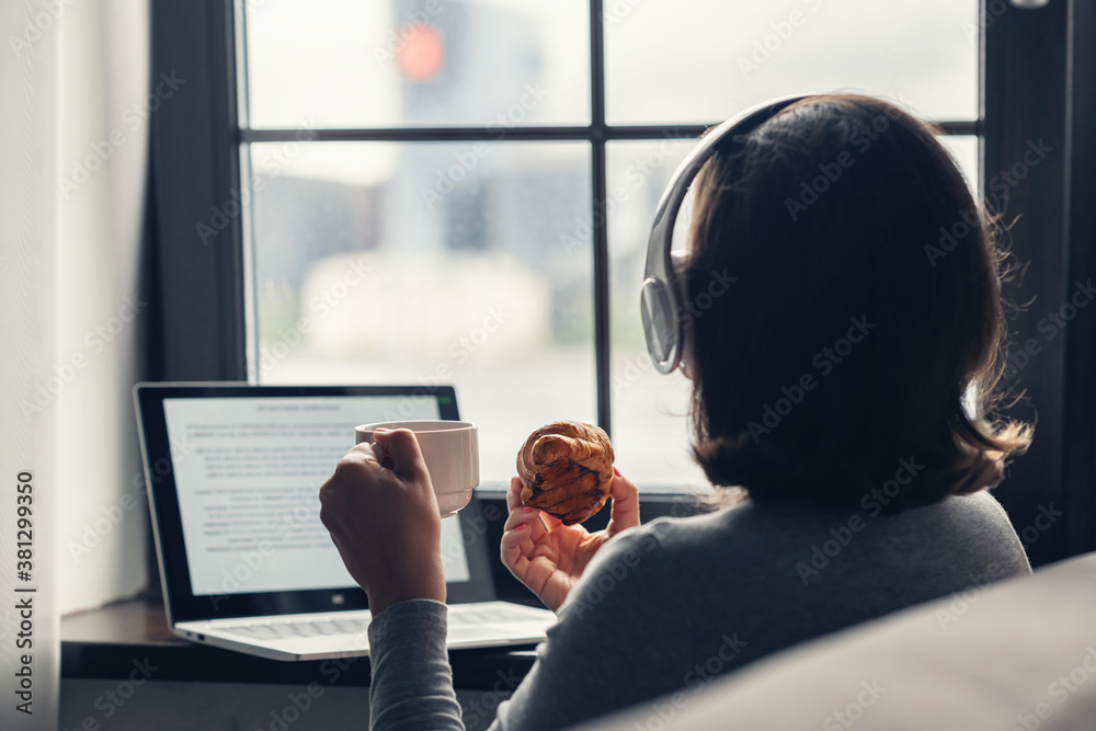 Back view of lonely woman enjoying having breakfast with cup of coffee and croissant, listenning music in headphones working on laptop sitting near window in cafe. - obrazy, fototapety, plakaty 