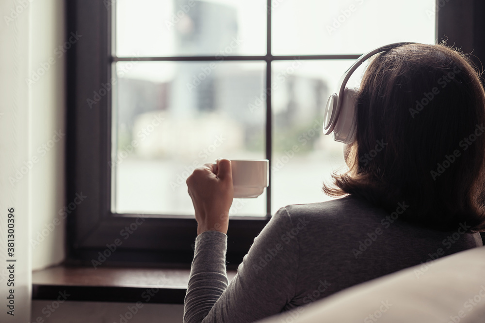 Φωτογραφία Back view of lonely сaucasian woman  with cup of coffee, listenning music in headphones sitting near window at cold winter morning