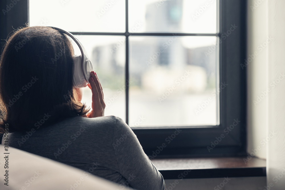 Back view of сaucasian young woman in headphones enjoying listening music sitting near window of apartments. Back view of сaucasian young woman in headphones enjoying listening music sitting near window of apartments.