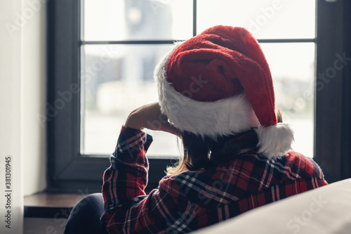 Back view of lonely sad young woman in a red santa claus christmas hat sitting and looking at window.