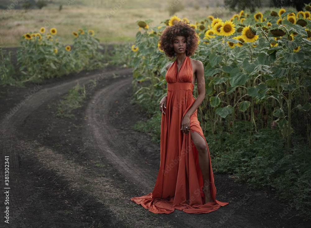 Young black woman in a long orange dress walks in the sunflower field.