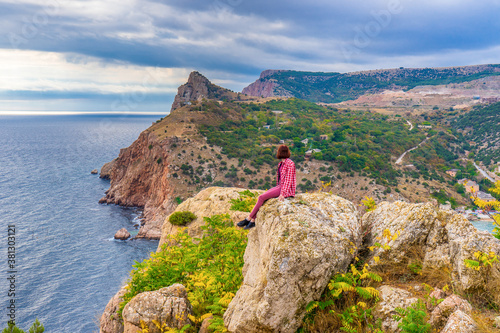 Teen tourist girl sitting on mountain cliff against beautiful seascape in cloudy day. Young lady relaxing on background of sea, mountains and sky. Travelling and discovering distant places of Earth