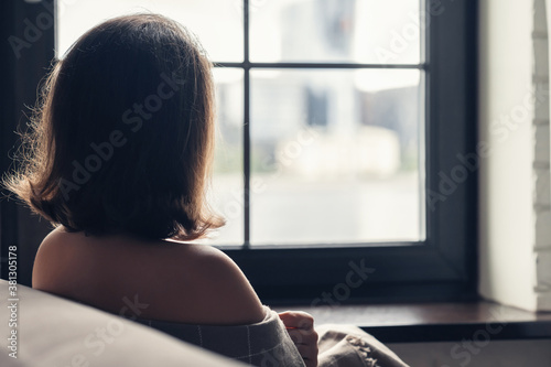 Back view of lonely сaucasian young woman wrapped in a plaid enjoying sitting near window of apartments.