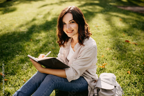 Young woman student sitting on grass in a park with notebook, writing, looking at camera.