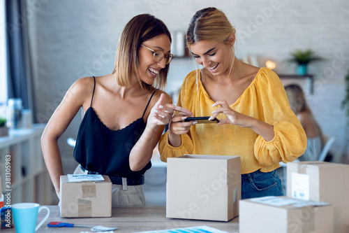 Two beautiful freelance business women using smart phone to taking photos to sell online while preparing pack product box in her startup small business. photo