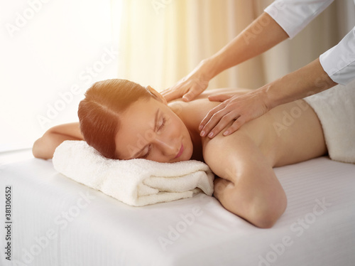 Pretty brunette woman enjoying procedure of back massage in sunny spa salon. Beauty concept